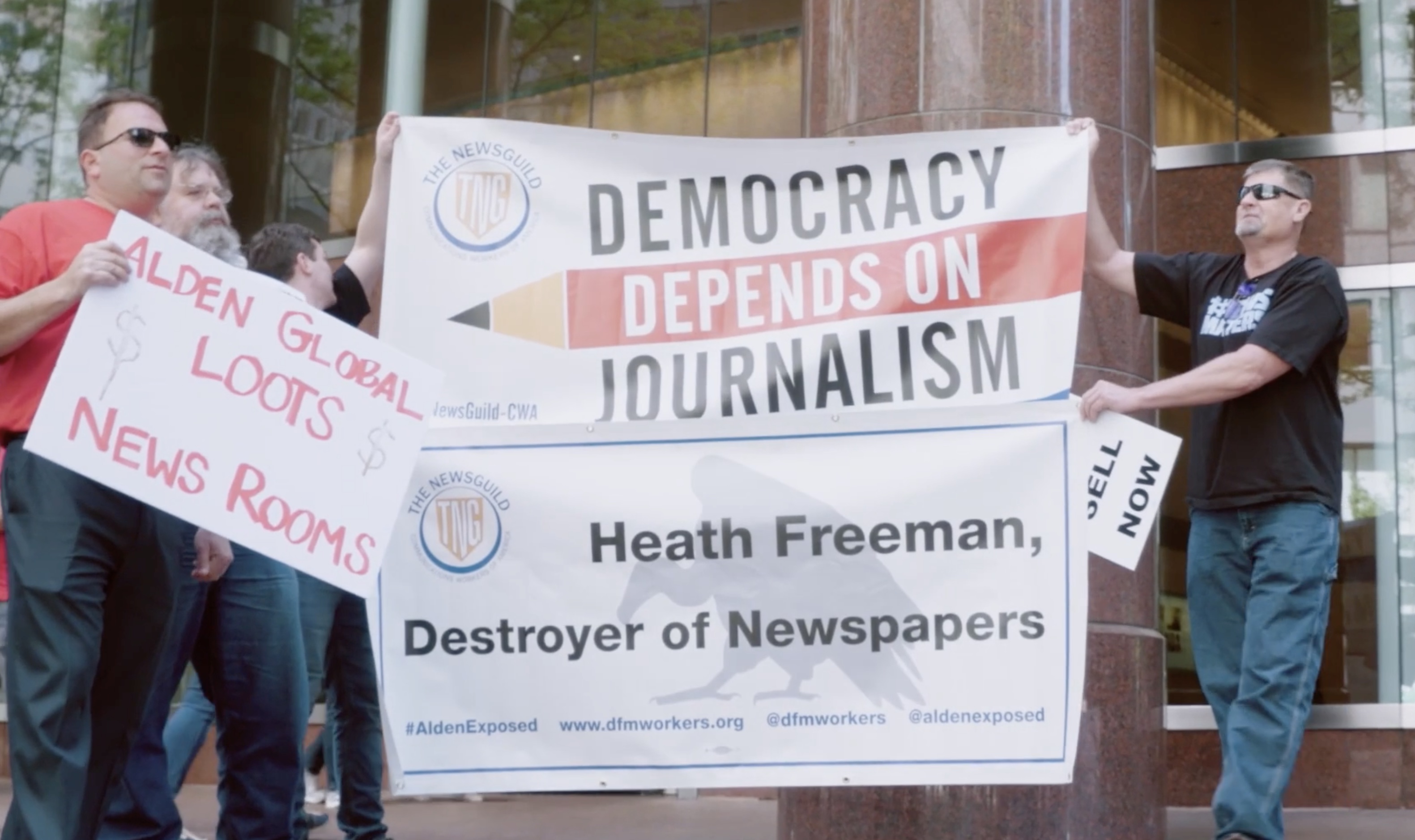 A still image from Stripped From Parts of journalists protesting. Two hold up a large sign with "Democracy Depends on Journalism" and "Heath Freeman, Destroyer of Newspapers."