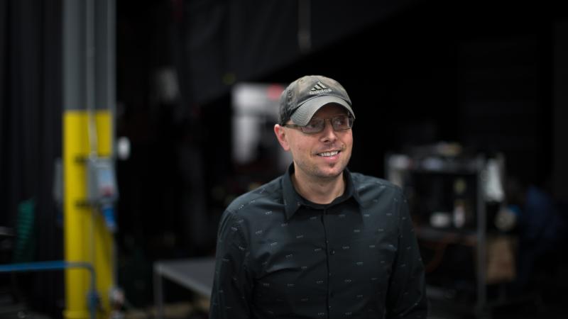 A medium shot of filmmaker Adrian Prawica, who is wearing a black button down shirt and a baseball cap. He is looking off to the right.