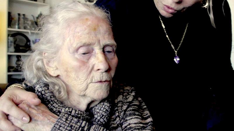 A care worker looks down at an elder woman. They hold hands, and the elder woman's eyes are downcast.