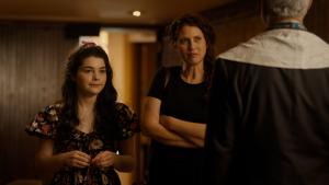 A mother with brown hair, wearing black and her young daughter who is non-verbal and disabled wearing a dress with a flower print, confronts the rabbi who we only see the back of in the synagogue sanctuary. 