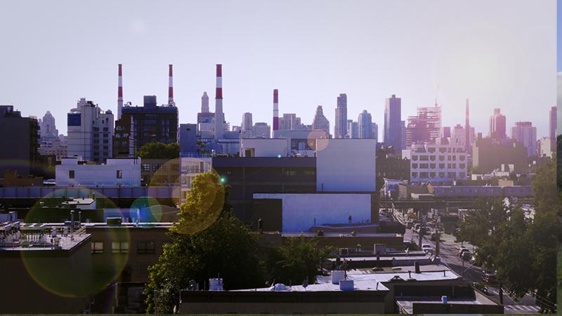Wide angle shot of a city showing power plant chimneys and an elevated train line from the film How To Power A City