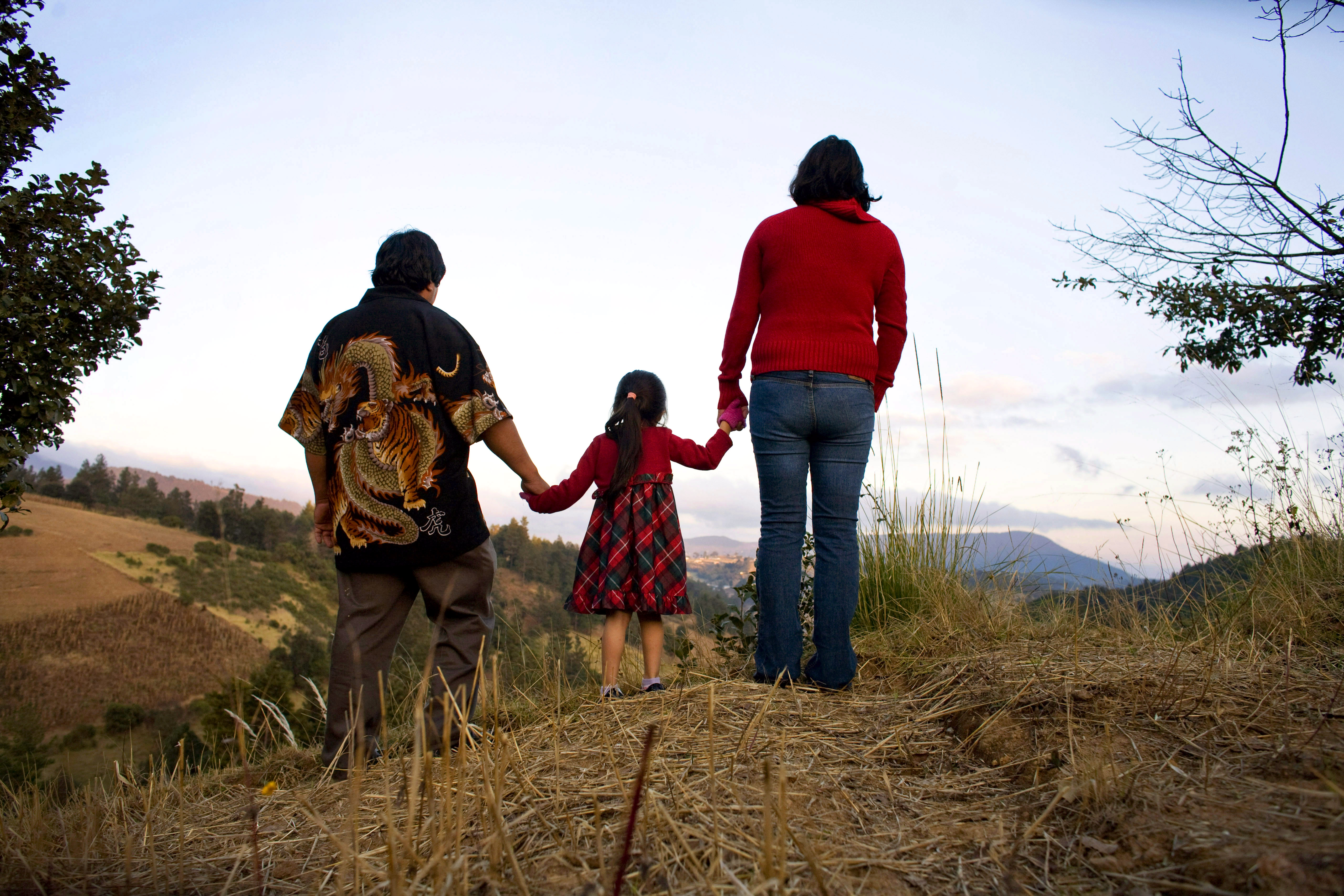 A still from the New Day film Sin Pais. The backs of three family members holding hands, a small child in the middle. They walk on a sandy path with tall grass on either side.