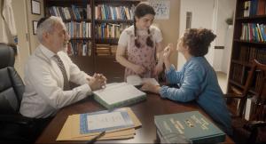 A mother (wearing a jean jacket, brown hair) sits and her daughter (non-verbal, disabled, brown hair in braids, wearing a white shirt and pink jumper) stand next to a desk in the rabbi's office. He sits across from them wearing a white shirt and tie, and black kippah. 