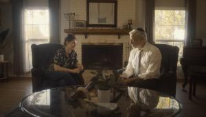 In a living room where we can see Judaica on a fireplace mantle (menorah) and on the glass table (shofar) sits the rabbi (white hair, black kippah, white shirt) and Leah (hair up, flower shirt and jeans). 
