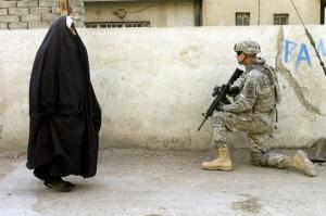 An Iraqi woman wearing a black burqa walks past a kneeling, armed American soldier