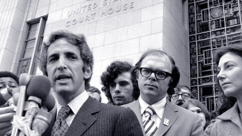 A black and white still image from the film, The Most Dangerous Man in America. The camera looks up from below at Daniel Ellsberg, a white man in a pinstriped suit, who speaks into a mass of TV reporter microphones that are taped together. He stands in front of tall, pale building with “United States Court House” embossed on the exterior wall. He is surrounded by men of various ethnicities who listen on or jockey into position to get their microphone closer to him.