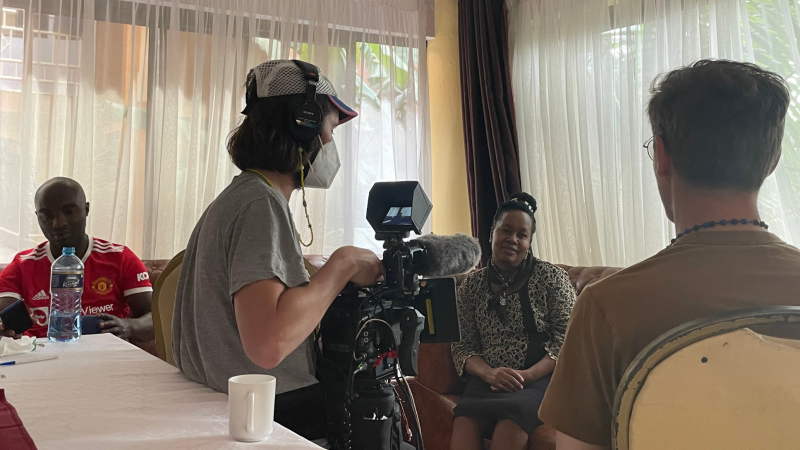 Production still for the film “Rollout.”  Filmmaker Trevor Zimmer and his cinematographer, who are both white, film an interview with a Kenyan woman.  The cinematographer wears a face mask over his nose and mouth.  On the left, a Kenyan man looks at his phone.  He sits outside of the cinematographer’s shot and is not part of the filmed interview.